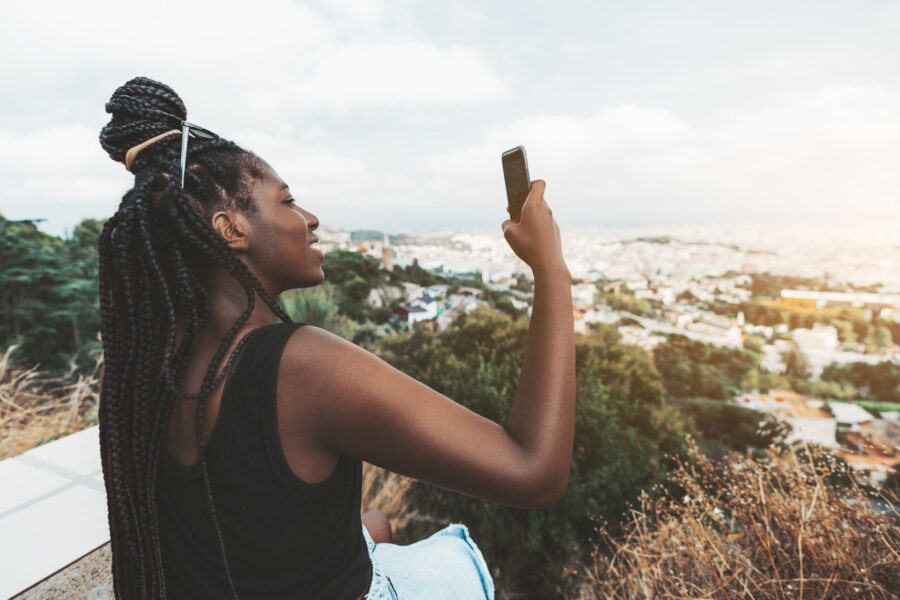 A cheerful cute African girl is photographing amazing evening cityscape from high above using her smartphone; young black female with braided hair using the cellphone to take pictures of a landscape