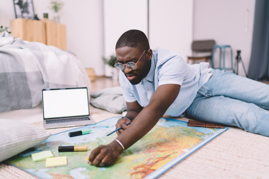 African American man in casual clothes looking for interesting places on map and making notes while sitting on floor near laptop at home