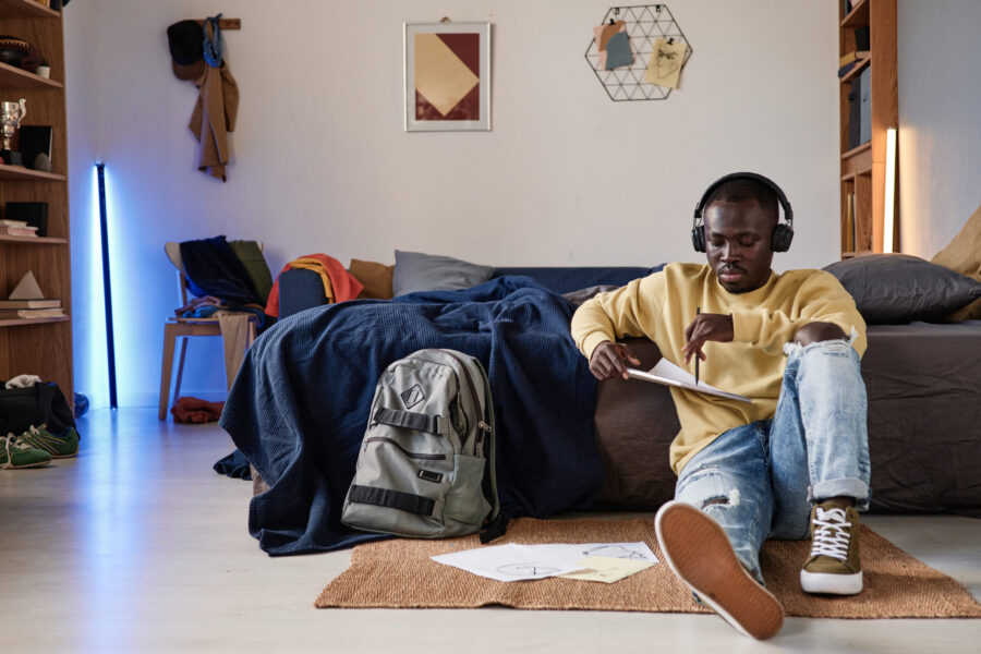 Serious African-American student guy in headphones sitting on floor in room with neon sticks and drawing illustration in sketchpad